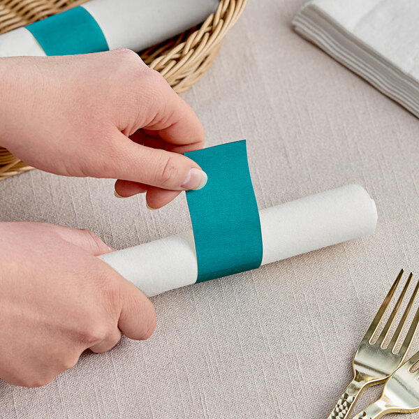 A person's hands wrapping a teal paper napkin band around a napkin.